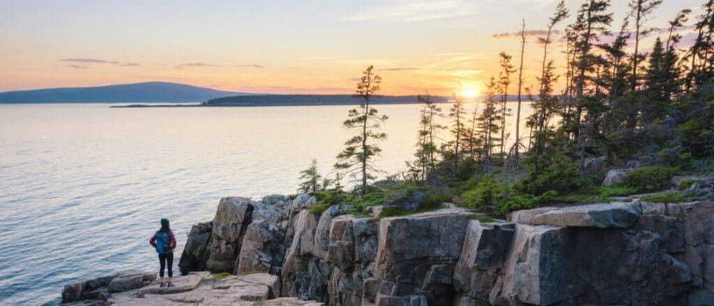 A person stands on a rocky cliff overlooking a serene body of water at sunset. The sky is painted with warm hues, and the silhouette of trees lines the edge of the cliff. A distant mountain is visible across the water.
