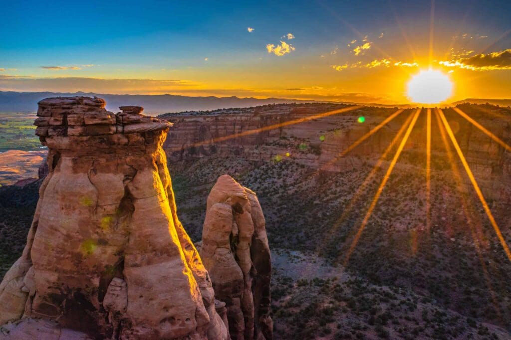 Sunset over a rugged canyon landscape with dramatic rock formations in the foreground. The sky is a vibrant mix of orange and blue hues as the suns rays spread across the horizon, casting warm light on the rocky terrain below.