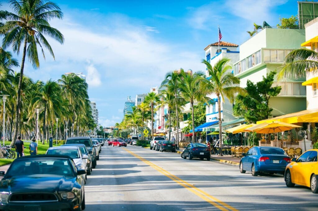 A lively street with cars parked along both sides, lined with palm trees and colorful buildings. Outdoor cafes have yellow umbrellas, and the sky is bright blue with scattered clouds. Pedestrians stroll on the sidewalk.