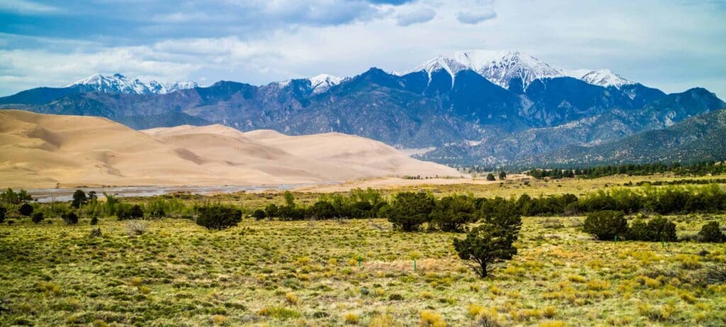 A landscape featuring rolling sand dunes in the foreground, grassy plains with scattered trees, and a backdrop of snow-capped mountains under a partly cloudy sky.