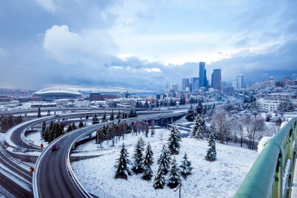 Snow-covered landscape featuring a highway with light traffic, leading to a city skyline. Evergreen trees dot the white ground, and a large stadium is visible to the left. The sky is overcast with patches of blue.