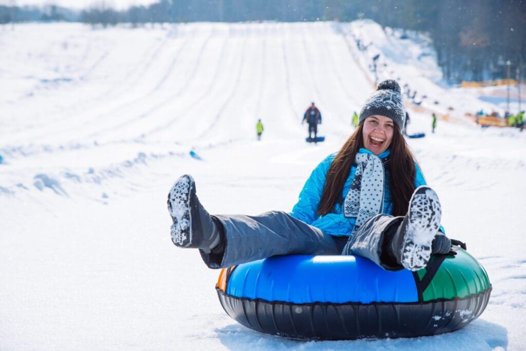 A person in a blue jacket and knit hat is joyfully snow tubing down a snowy hill, with feet up in the air. Snow-covered slopes and other people in the background create a lively winter scene.