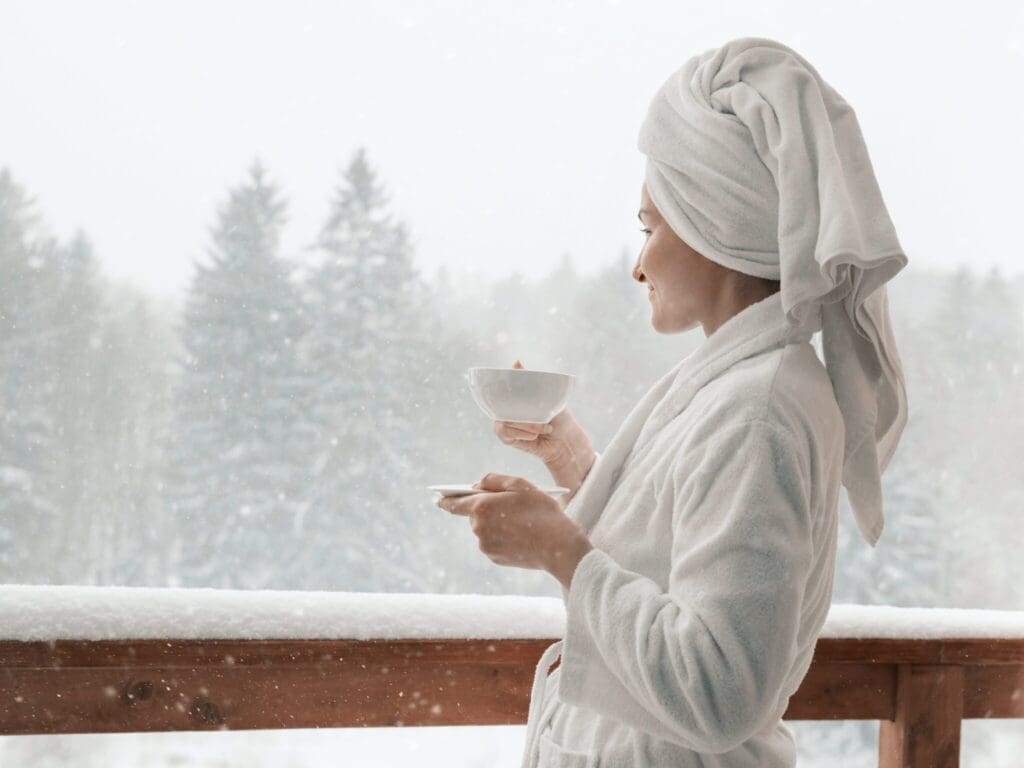 A person in a white robe and towel wrapped around their head stands on a snowy balcony, holding a cup and saucer. They are looking out at the snow-covered trees in the background, enjoying a peaceful winter scene.