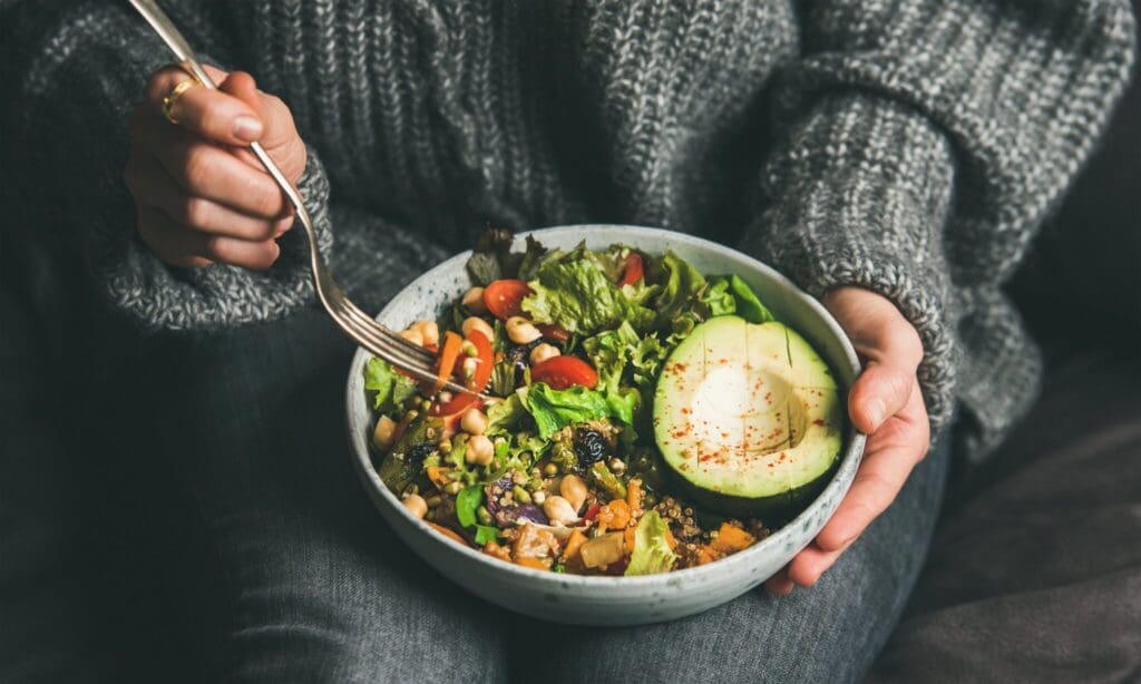 A person in a gray sweater holding a bowl of salad on their lap. The salad includes cherry tomatoes, avocado, beans, lettuce, and other vegetables. The fork is poised above the bowl, about to pick up some salad.