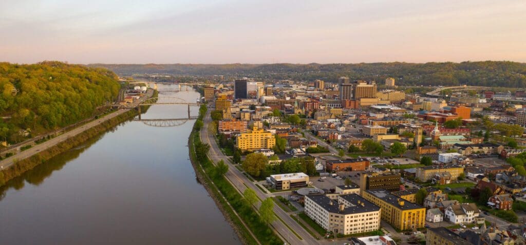 Aerial view of a city at sunset with a river running alongside. The skyline features tall buildings and bridges over the river. Greenery lines the riverbank, and the cityscape is dotted with a mix of commercial and residential buildings.