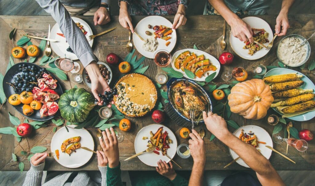 Top view of a festive table with various dishes, including roasted vegetables, corn, a pumpkin pie, grapes, and a roasted turkey. People are serving themselves and reaching across the table, surrounded by autumn leaves and decorative pumpkins.