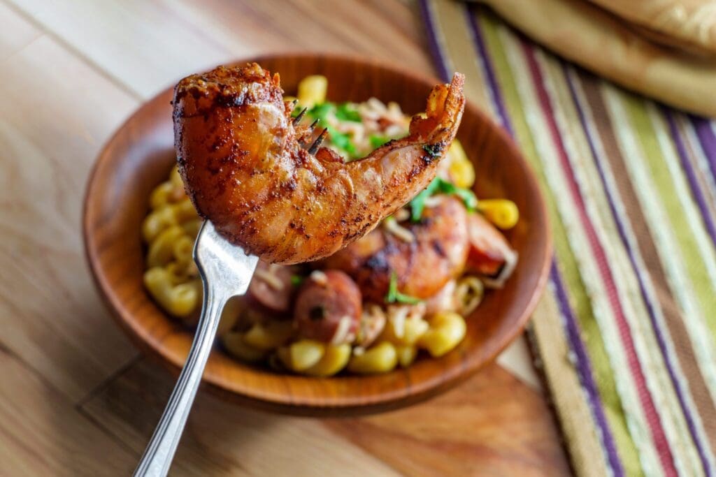 A fork holds a grilled shrimp above a wooden bowl filled with fusilli pasta, sliced sausage, and herbs. The dish is placed on a wooden surface with a striped cloth beside it.