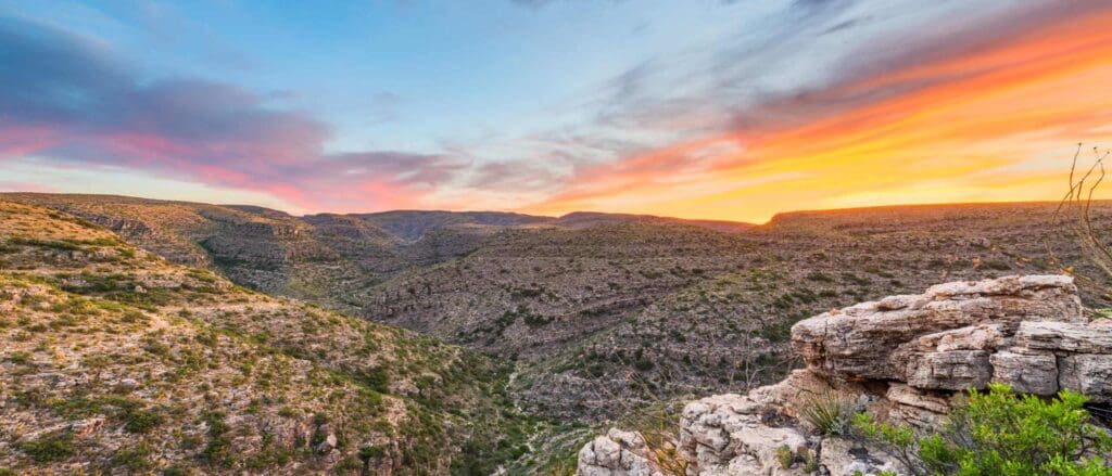 View of a rugged canyon at sunset with layered rock formations and sparse vegetation. The sky is vibrant with hues of orange, pink, and blue, casting a warm glow over the landscape.