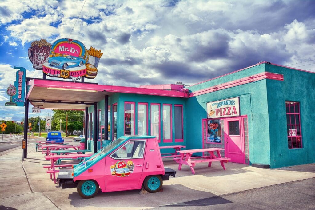 A brightly colored retro diner with a Route 66 theme. The building is turquoise with pink trim, featuring a vintage-style sign for Mr. Dz Diner. A small, pink vintage car is parked in front. Cloudy sky in the background.