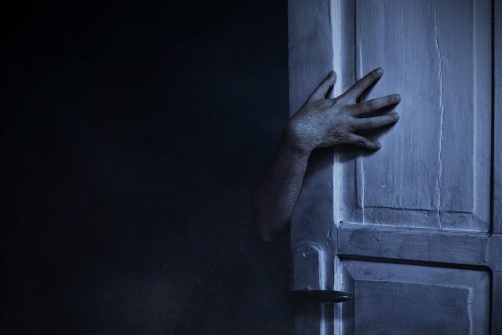 A dimly lit scene showing a partially open wooden door with a hand reaching out from the darkness beyond. The hand grips the door edge, casting an eerie and mysterious atmosphere. The background is shadowy and undefined.