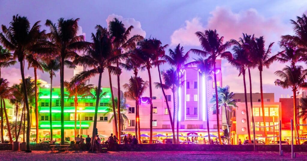 Palm trees silhouette against a vibrant, neon-lit street at dusk. Colorful lights illuminate art deco buildings, creating a lively atmosphere. People gather in front, enjoying the scene. The sky is a mix of purple and pink hues.