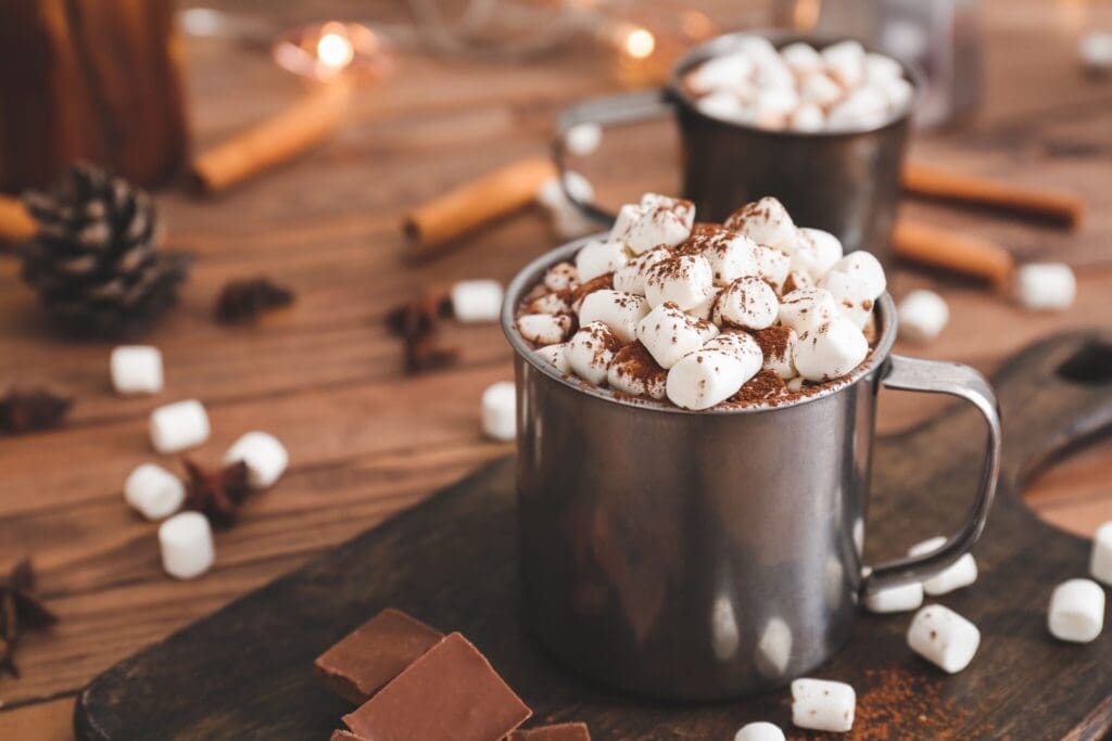Two metal mugs filled with hot chocolate topped with marshmallows and cocoa powder sit on a wooden tray. Chocolate pieces are nearby, with cinnamon sticks and pine cones in the blurred background, creating a cozy atmosphere.