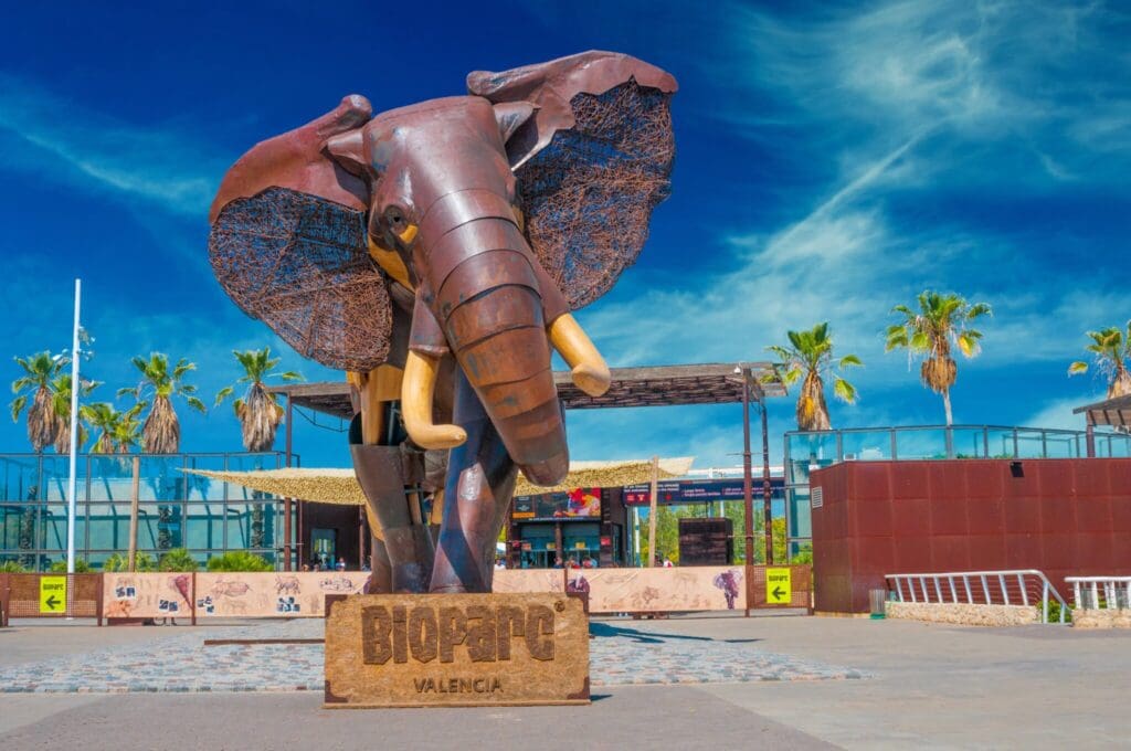 A large, stylized elephant sculpture stands at the entrance of Bioparc Valencia. The artwork is made of metal with detailed texture on the ears and tusks. Palm trees and a clear blue sky are in the background, with signage directing visitors.