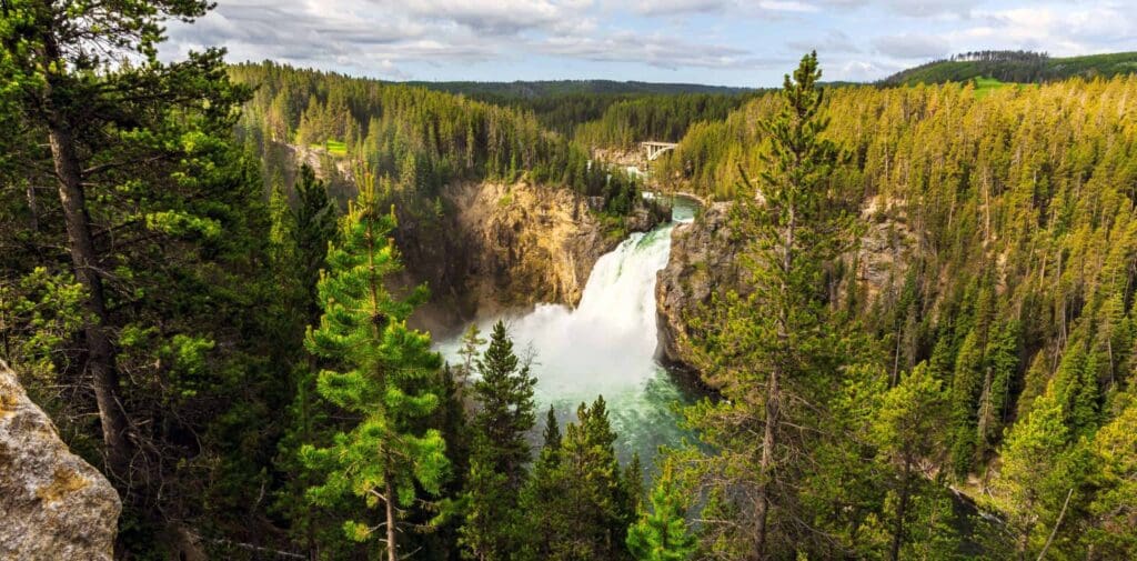 A scenic waterfall cascades into a river surrounded by dense pine forests under a partly cloudy sky. A bridge is visible in the distance amidst the greenery.
