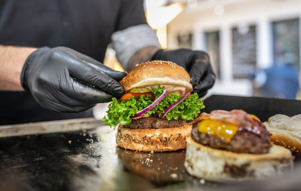A person wearing black gloves assembles a hamburger, adding lettuce, tomato, onion, and a sesame seed bun on a beef patty on a grill. Another burger with bacon and cheese is partially visible beside it.