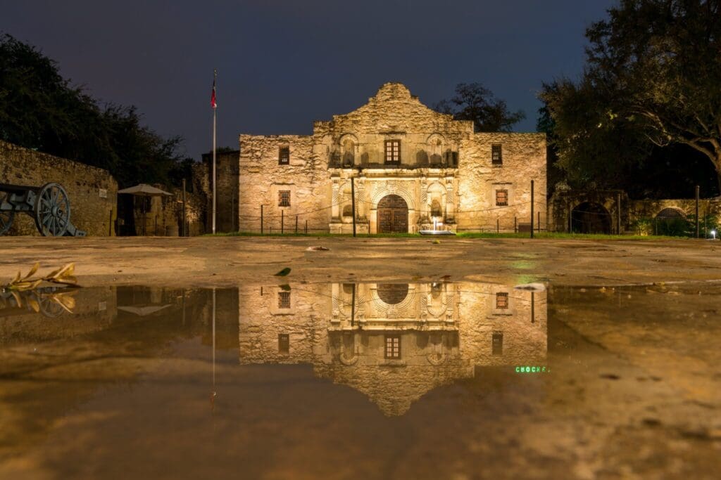 The Alamo at night with its façade illuminated, reflecting in a puddle in the foreground. A flagpole with a flag stands to the left, and a cannon is visible near the trees on the left. The sky is dark, and the scene is tranquil.