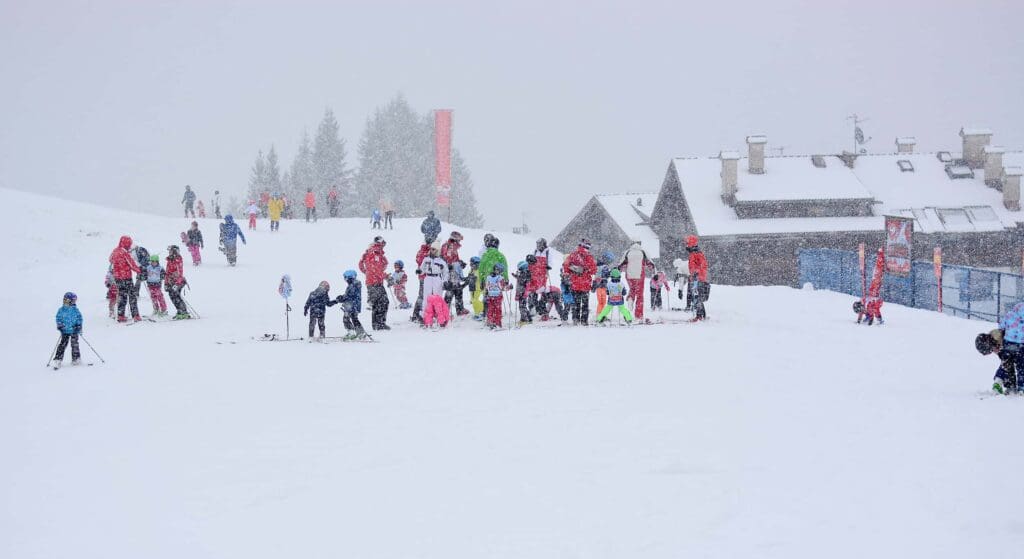 A snowy ski slope with a group of people in colorful ski attire gathered near a building. Snowflakes are falling, partially obscuring the view. Other skiers are visible in the background, amidst snow-covered trees.