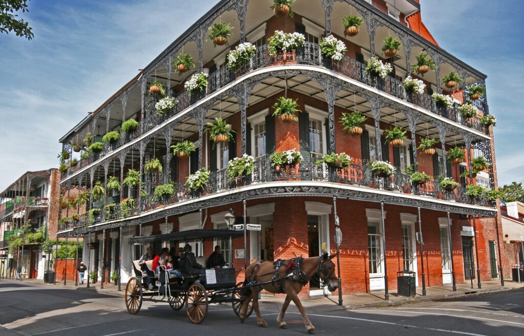 A historic red brick building features ornate black iron balconies adorned with hanging plants and flowers. A horse-drawn carriage with several passengers passes by on the street in front. The scene is sunny and vibrant.