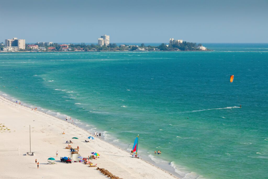 Aerial view of a beach with white sand and turquoise water. People are lounging under colorful umbrellas along the shoreline. A kite surfer is visible in the water. Buildings and trees line the distant coastline under a clear blue sky.