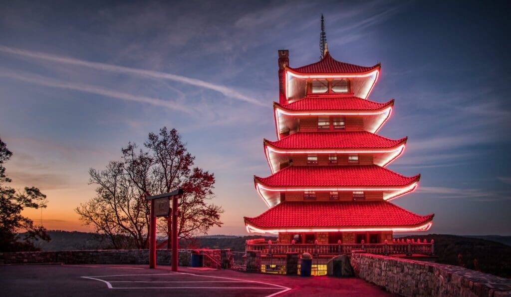 A five-story pagoda with red illuminated edges stands against a twilight sky. Trees and a stone railing surround the area, and the structure overlooks a scenic view with a hint of sunset on the horizon.