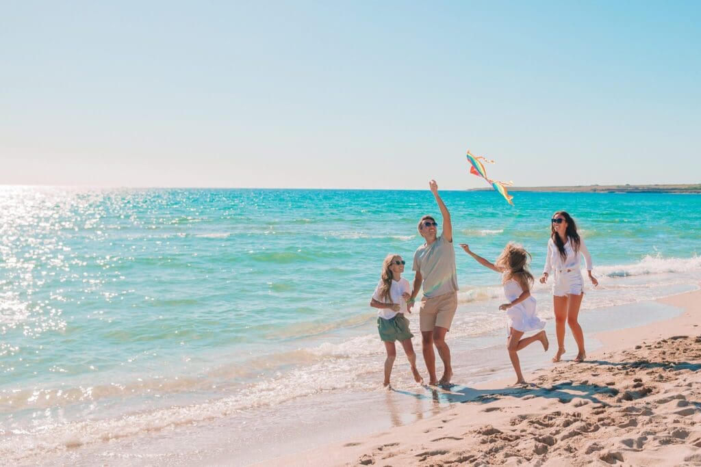 A family of four enjoys a sunny day at the beach. Two adults and two children, one holding a kite, walk along the shoreline with gentle waves in the background. The children are smiling and running on the sandy beach.