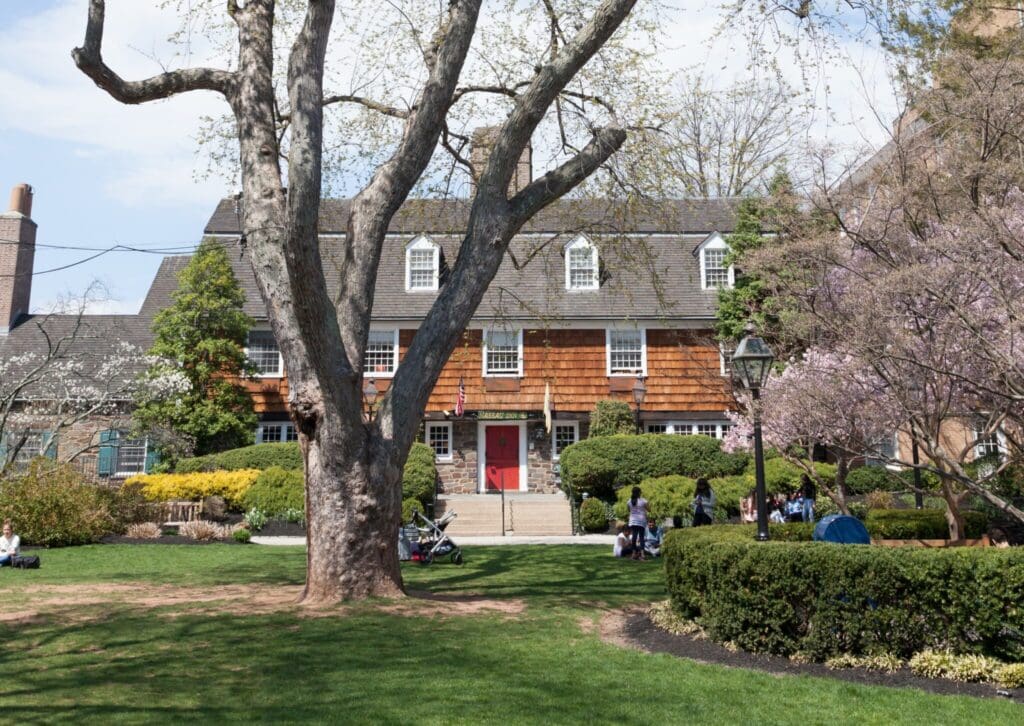 A large, two-story brick building with a sloped roof and numerous windows is surrounded by lush greenery. A prominent tree stands in the foreground, and people are seated on the lawn enjoying the sunny day.