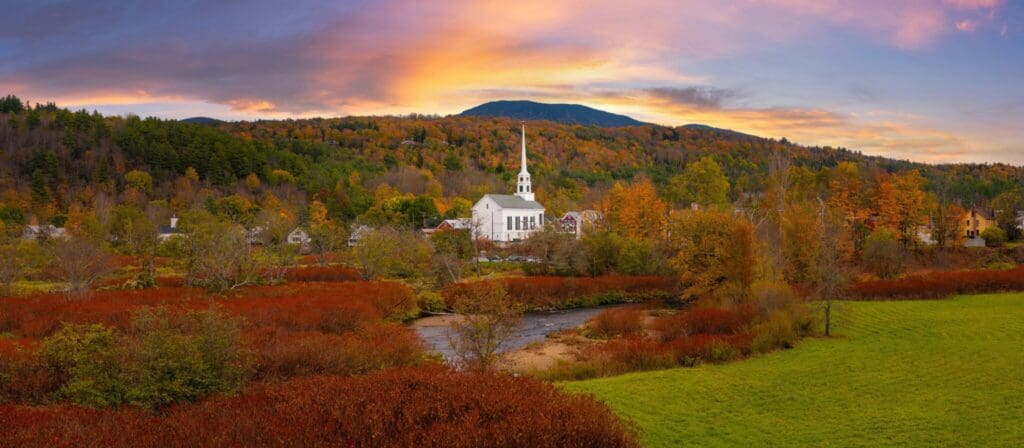 A picturesque landscape featuring a quaint village with a white church, surrounded by vibrant autumn foliage. A gentle stream flows through the scene, set against rolling hills and a colorful sunset sky.