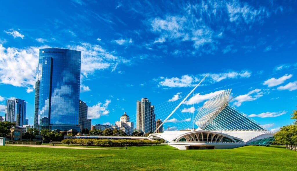 Milwaukee skyline featuring modern buildings, including a distinctive white, winged structure. This structure is set against a bright blue sky with scattered clouds, surrounded by green grass in the foreground.