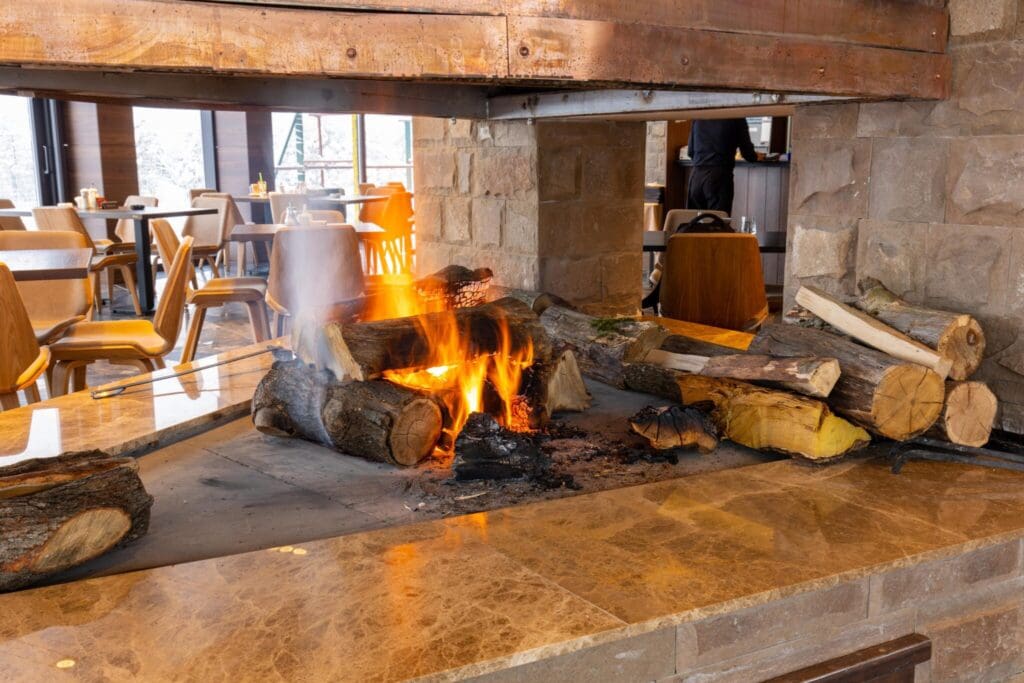 A cozy indoor wooden fire burns on a stone hearth within a restaurant. Wooden logs are stacked on the fire. In the background, tables and chairs are arranged, and a person stands near a window.