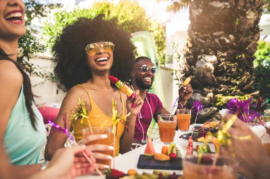 Group of friends enjoying an outdoor party, laughing and holding colorful drinks. A variety of fruit platters are on the table. Palm trees and sunlight create a relaxed, festive atmosphere.