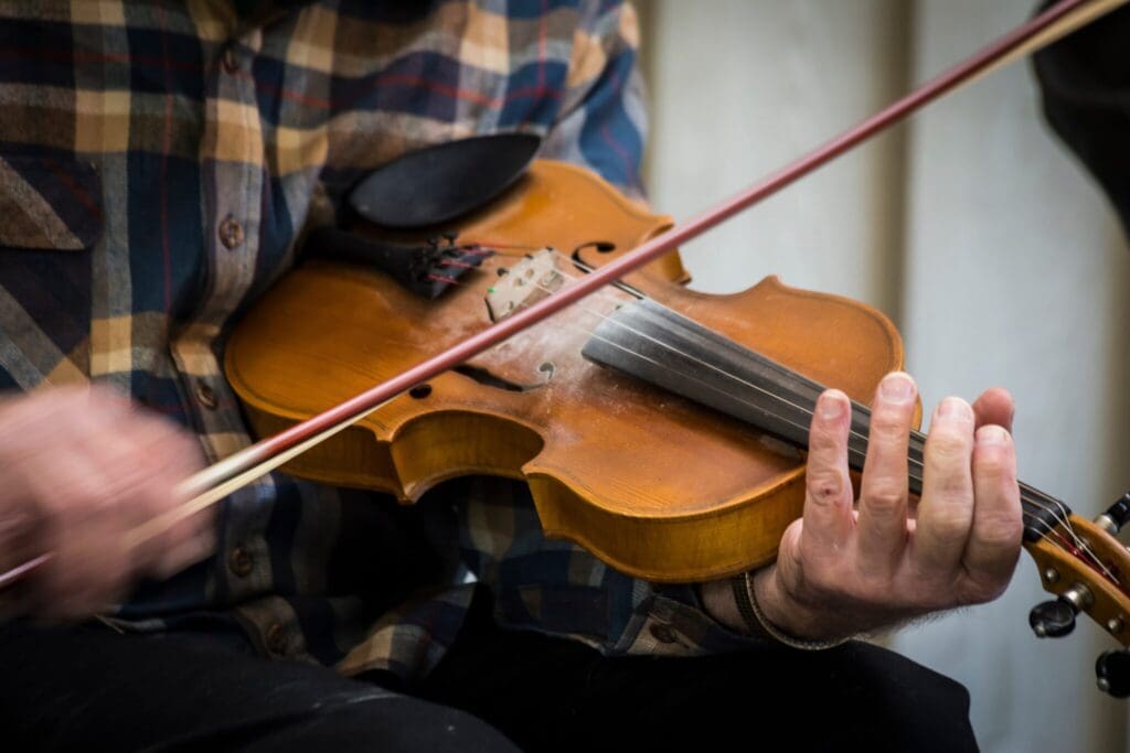 A person wearing a plaid shirt plays a violin, focusing on the strings with a bow. The image shows a close-up of the hands and the instrument, emphasizing the skill and detail involved in playing.