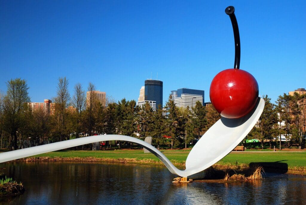A large sculpture of a spoon with a cherry on top stretches across a pond in a park. Trees line the background, and several tall buildings are visible under a clear blue sky.
