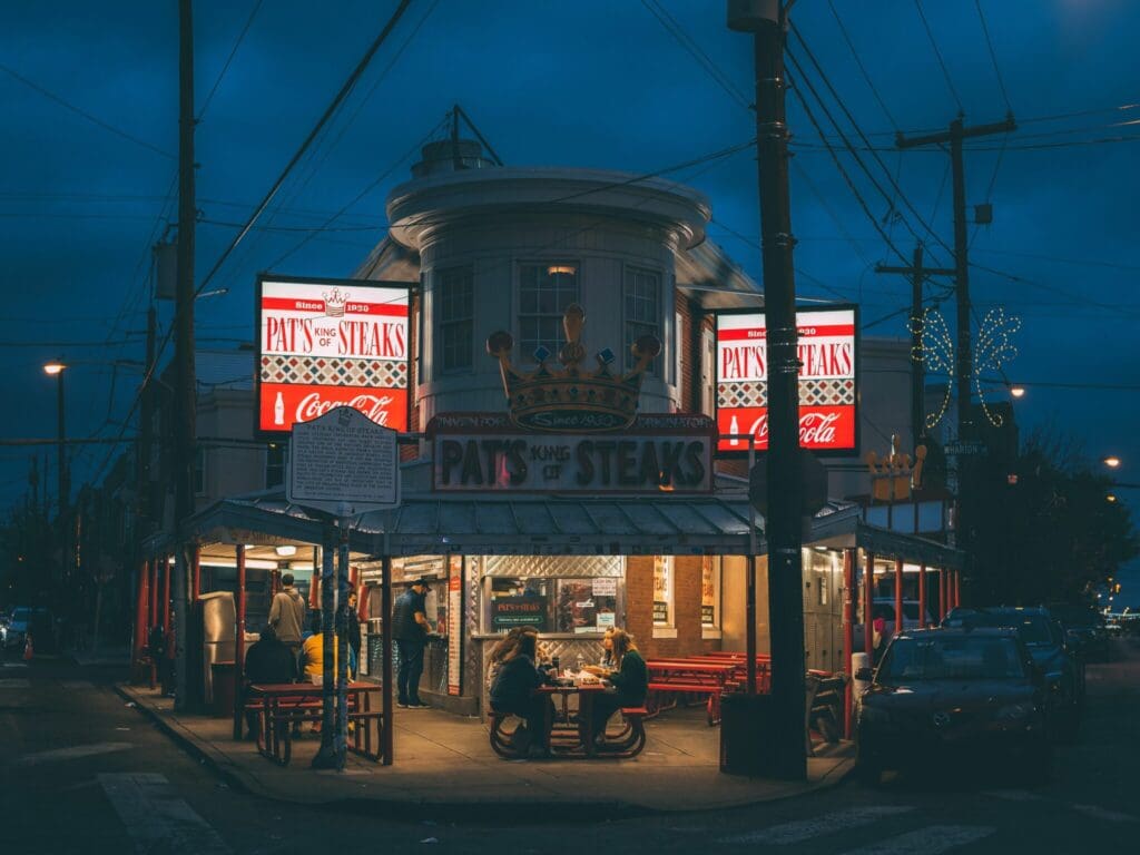 A night scene at Pats Steaks, a corner eatery with bright red and white signage. A lit interior reveals patrons sitting at outdoor tables under a roof. Streetlights illuminate the area, and an overcast sky sets a moody atmosphere.