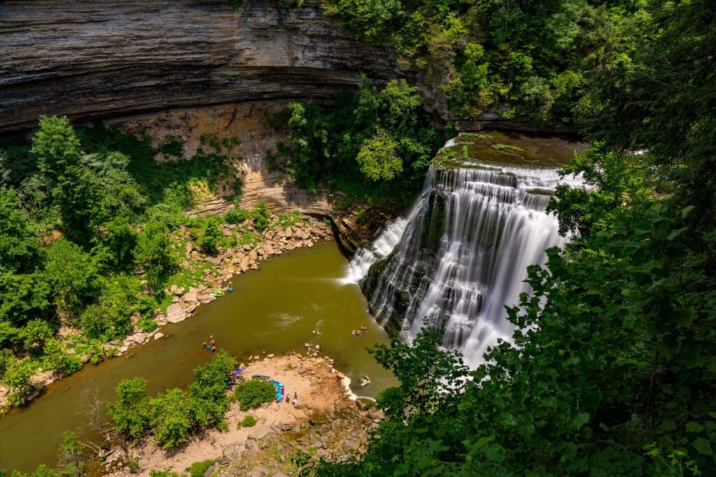 Aerial view of a waterfall cascading into a green pool surrounded by rocky cliffs and lush greenery. People are visible on a sandy area near the waters edge, basking in sunlight. The scene is vibrant and natural.