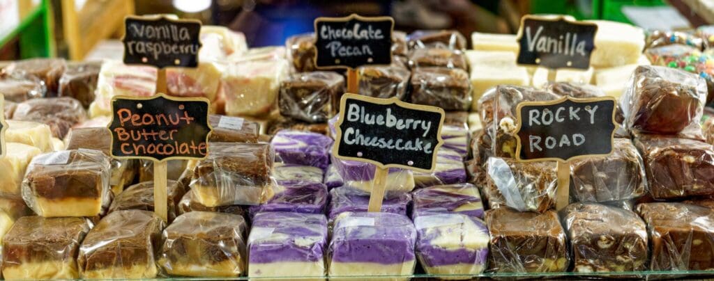A display of various fudge flavors wrapped in plastic, including peanut butter chocolate, blueberry cheesecake, rocky road, chocolate pecan, vanilla raspberry, and vanilla. Small chalkboard signs label each flavor.