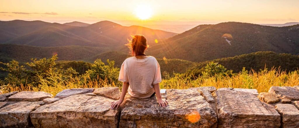 A person sits on a stone wall, gazing at a sunset over a mountainous landscape. The scene features vibrant greenery and a bright orange sky with the sun setting behind distant hills. The persons hair is gently blowing in the breeze.