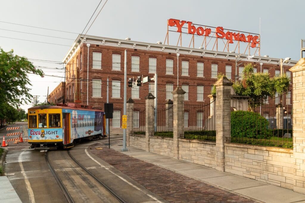 A colorful tram with a Coors Light advertisement travels down tracks beside a brick building labeled Ybor Square. The weather is overcast, and the scene includes street fixtures like signs and barriers.