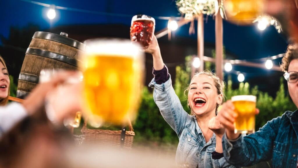 A group of people raising glasses of beer in an outdoor setting at night. String lights and a large wooden barrel are in the background. They appear to be celebrating or toasting, with a woman at the center laughing joyfully.