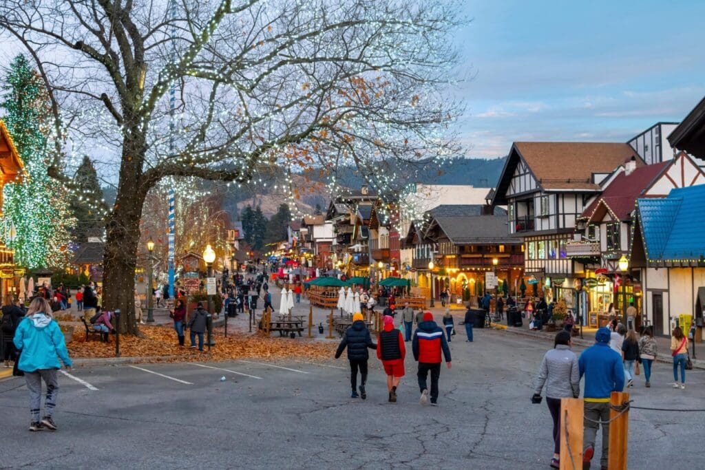 A festive street scene with people walking along a decorated town with Christmas lights and holiday decorations. Buildings resemble alpine architecture. A large tree with lights is on the left, and the atmosphere is lively and cheerful.