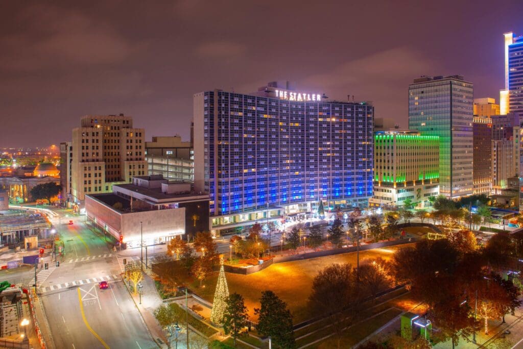 Night view of a cityscape featuring a large illuminated building with The Statler sign. Streets surround the building, displaying urban lighting and light traffic. Trees and a small park area are visible in the foreground.
