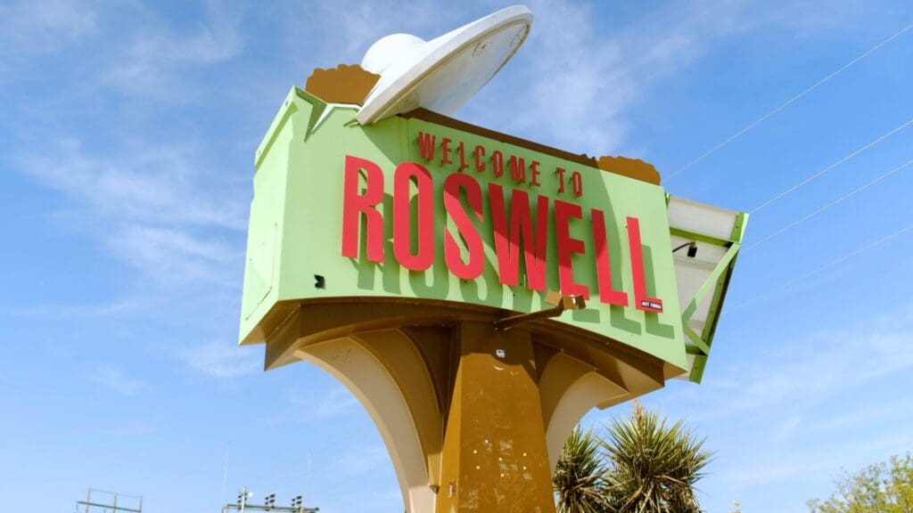 A large, retro-style sign reads “Welcome to Roswell” in bold red letters. The sign has a UFO design element on top and is set against a clear blue sky with a few clouds. The base of the sign is a light brown color.