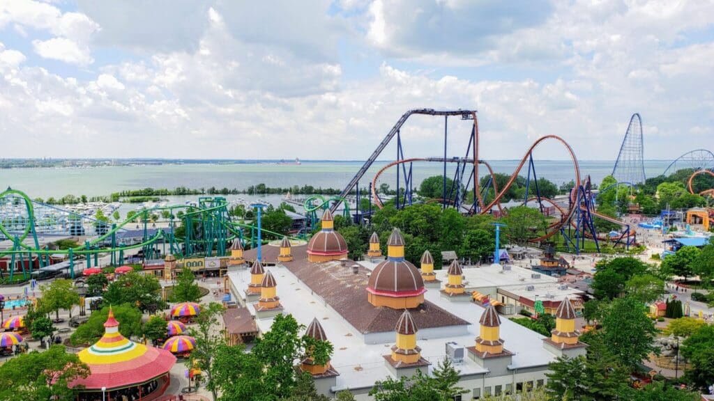 Aerial view of an amusement park with various colorful roller coasters and rides. A large lake and cloudy sky are in the background. The foreground features buildings with decorative domes and vibrant tents. Lush greenery surrounds the area.