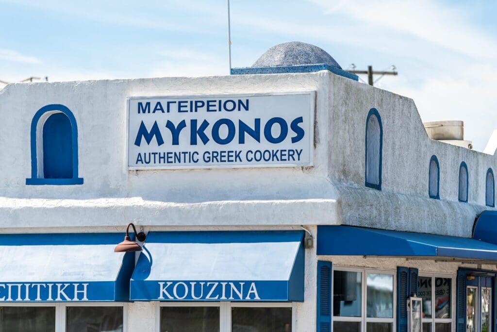 A bright white and blue building with a sign reading Mykonos Authentic Greek Cookery in both Greek and English. The structure features traditional Greek architectural elements, like rounded edges and a small dome.