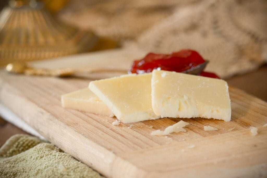 Slices of cheese on a wooden cutting board with a spoonful of red jam in the background. A textured cloth and a hint of a golden object are partially visible.