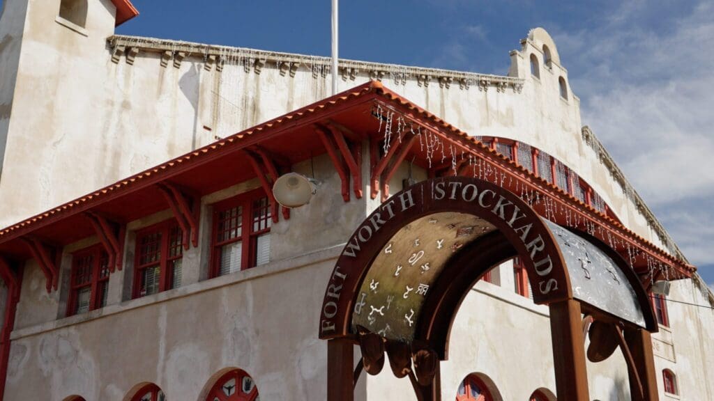 Historic building at Fort Worth Stockyards with a unique blend of red and beige architecture. A black wrought iron archway in the foreground displays the words Fort Worth Stockyards and is decorated with cattle brands. Blue sky above.
