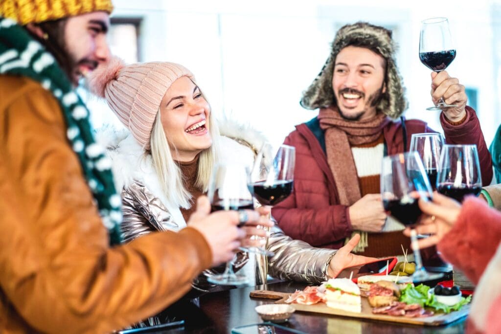 A group of people in winter clothing enjoy each others company, holding glasses of red wine. They are gathered around a table with a platter of assorted appetizers, laughing and toasting. The scene is warm and lively.