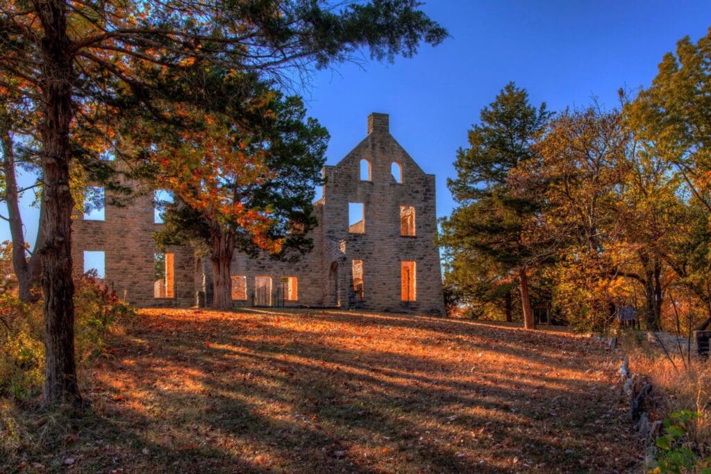 Stone ruins of a building stand amidst trees with vibrant autumn foliage. The sunlight casts long shadows across the fallen leaves on the ground, creating a warm, serene atmosphere under a clear blue sky.