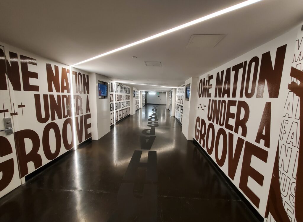 A hallway with white walls features large brown text that reads ONE NATION UNDER A GROOVE in bold, block letters. The phrase repeats on adjacent walls, and lighting from the ceiling illuminates the corridor and the reflective dark floor.