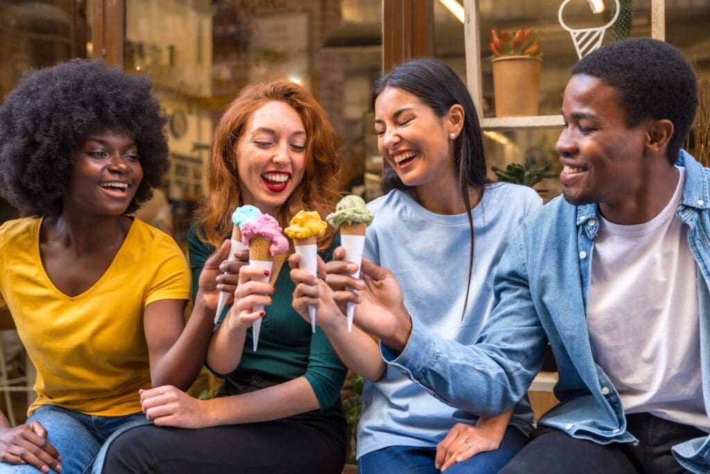 Four friends sit together, smiling and holding ice cream cones. They appear happy and are dressed casually. The background shows a shopfront with plants and decorative items.