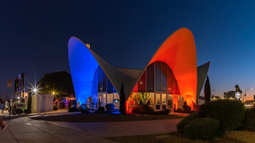 A futuristic building with curved, wing-like structures is illuminated at night in red and blue lights. The sky is a deep blue, and surrounding greenery is visible in the foreground.